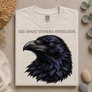 Puede incluir: Una camiseta de color crema con una ilustración detallada de la cabeza de un cuervo negro de perfil. Sobre el ave, el texto "SEE WHAT OTHERS OVERLOOK" aparece en letras mayúsculas grises. La camiseta tiene cuello redondo y una etiqueta de la marca Comfort Colors.