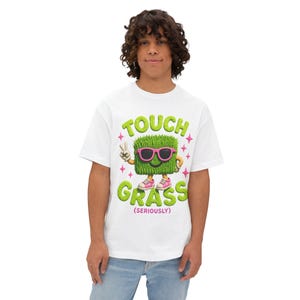 May include: White t-shirt featuring a cartoon graphic of a square of grass wearing sunglasses and sneakers. The text "TOUCH GRASS (SERIOUSLY)" is in green and pink. The shirt is worn by a person.