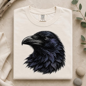 Puede incluir: Una camiseta de manga corta color crema con una ilustración gráfica detallada de una cabeza de cuervo negra de perfil. La camiseta tiene cuello redondo y una etiqueta de la marca Comfort Colors impresa en el cuello interior con la talla L-G-G.
