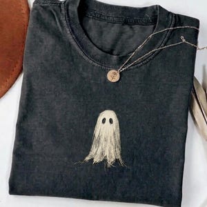 May include: A charcoal gray crew neck t-shirt featuring a central graphic of a beige, sketch-style ghost with hollow eyes. A delicate gold-toned chain necklace with a small circular pendant engraved with a rose design rests on the neckline.