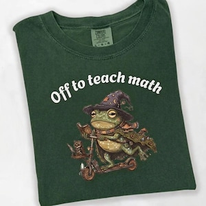 May include: Dark green t-shirt with the text "Off to teach math." The shirt features a whimsical illustration of a frog wearing a wizard hat and scarf, riding a scooter, with an owl companion.
