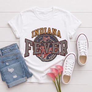 May include: White t-shirt with the Indiana Fever logo, featuring the text "INDIANA FEVER" in distressed lettering. The design includes a basketball and the text "WNBA". The shirt is paired with blue jeans and white sneakers.