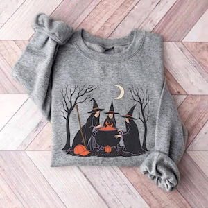 May include: A heather gray crewneck sweatshirt featuring a graphic illustration of three witches in black robes and pointed hats standing around a bubbling orange cauldron, with bare trees, a crescent moon, and two orange pumpkins in the scene.