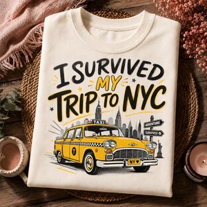 May include: A cream-colored t-shirt featuring a graphic of a yellow New York City taxi cab in front of a city skyline. The text reads I SURVIVED MY TRIP TO NYC. A signpost shows the words ADVENTURE and SURVIVED, with a small Statue of Liberty nearby.