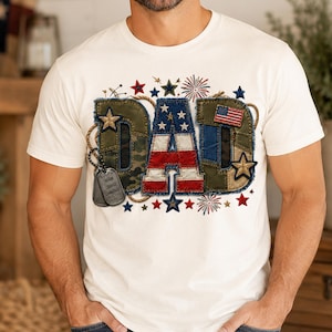 Könnte beinhalten: Ein weißes Kurzarm-T-Shirt mit dem Wort DAD in großen Blockbuchstaben. Die Buchstaben sind mit Mustern der amerikanischen Flagge, Tarnfarben und Denim-Texturen verziert. Das Design umfasst goldene Sterne, Feuerwerk und zwei Erkennungsmarken.