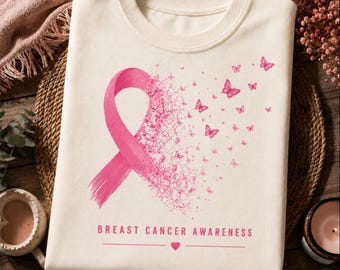 May include: A cream-colored t-shirt featuring a large pink awareness ribbon that transitions into a swarm of small pink butterflies. Below the graphic, the text reads BREAST CANCER AWARENESS with a small pink heart centered on a horizontal line.