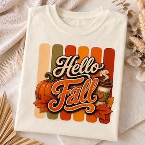 Puede incluir: Una camiseta color crema con un diseño gráfico de rayas verticales en tonos otoñales apagados. El texto "Hello Fall" está escrito en una fuente cursiva llamativa. El diseño incluye una calabaza naranja, una taza de café y hojas de arce.
