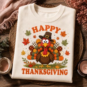 May include: A cream-colored t-shirt featuring a graphic of a turkey wearing a pilgrim hat, surrounded by colorful autumn leaves and the text "HAPPY THANKSGIVING" in orange letters. The turkey has a multi-colored tail with plaid patterns.