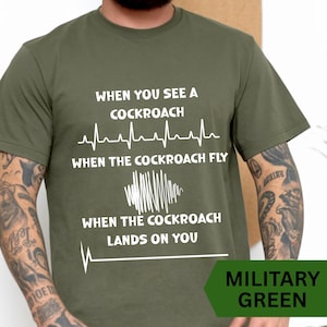 May include: A military green short-sleeved t-shirt featuring white text that reads: WHEN YOU SEE A COCKROACH, followed by a heartbeat line, WHEN THE COCKROACH FLY, a jagged sound wave graphic, WHEN THE COCKROACH LANDS ON YOU, and a flatline.
