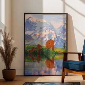 Puede incluir: Una impresión artística enmarcada representa un oso y su cachorro cerca de un lago con montañas al fondo. La obra de arte está en un marco negro y se exhibe en una habitación con una silla azul, una alfombra y una planta en maceta.