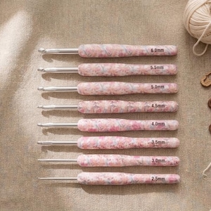 May include: A set of eight crochet hooks with silver metal tips and pink floral patterned handles. Each handle is clearly labeled with its size, ranging from 2.5mm to 6.0mm, indicating the specific diameter of the hook for various yarn projects.