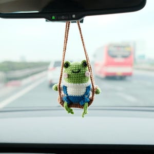 May include: A crocheted green frog figurine wearing blue overalls and a white shirt, sitting on a small brown swing suspended by brown yarn. The frog has black eyes and pink cheeks, hanging from a car rearview mirror above a digital display.