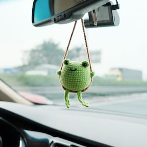 May include: A small, round, green crocheted frog ornament hanging from a brown string. The frog has two black eyes, a stitched smile, and dangling legs. It is suspended from the rearview mirror inside a vehicle, featuring a textured yarn surface.
