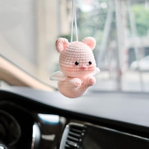 May include: A small, light pink crocheted pig ornament hanging from a white string. The pig features two small white wings on its back, two black bead eyes, and a tiny pink snout. The surface of the pig is textured with visible yarn stitches.