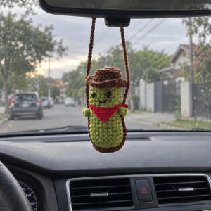 May include: A crocheted pickle ornament hanging from a rearview mirror. The green pickle wears a brown cowboy hat and a red bandana around its neck. It features two small black eyes and a stitched smile, suspended by a brown crocheted cord.