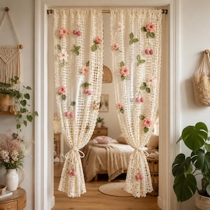 May include: Cream-colored crochet curtains with a grid pattern, adorned with pink flowers, green leaves, and pink cherry accents. The curtains are tied back with matching yarn and hang in a doorway, creating a soft, decorative room divider.