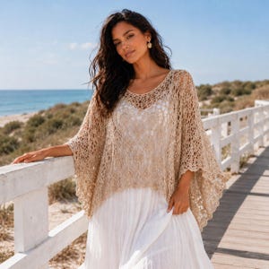 Könnte beinhalten: Ein beigefarbenes Poncho-Oberteil mit offenem Gewebe, einem Rundhalsausschnitt und weiten Ärmeln. Das Oberteil wird über einem weißen Kleid getragen. Der Hintergrund zeigt eine Strandszene mit einem Holzsteg und dem Meer.