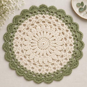 May include: A round, cream-colored crochet doily featuring a central floral-inspired pattern with intricate stitching. The doily is bordered by a wide, scalloped edge in a muted sage green yarn, creating a distinct contrast between the two colors.