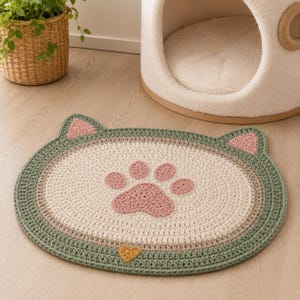 May include: Crocheted cat-shaped mat in cream, green, and pink. The mat features a paw print design in the center and cat ears at the top. A small yellow heart is at the bottom. The mat is designed for pets.