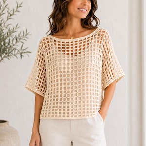 May include: A cream-colored, short-sleeved crochet top featuring a wide, open-grid square pattern. The garment has a boat neckline and a loose, boxy fit, worn over a solid, matching cream-colored camisole underneath, paired with light-colored trousers.