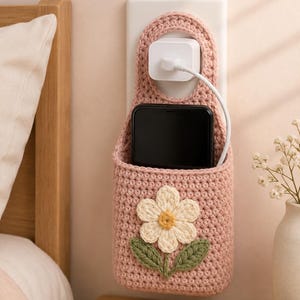 以下が含まれることがあります： ピンク色の、かぎ針編みで作られた壁掛け式の携帯電話ホルダー。デイジーのアップリケ付き。コンセントの上に吊るすように設計されており、中に携帯電話と充電器が入っています。デイジーはクリーム色の花びらと黄色の中心部、緑の葉が付いています。