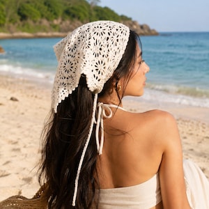 Puede incluir: Un pañuelo de cabeza de ganchillo color crema con un estampado floral. El pañuelo se ata en la parte posterior con largas cuerdas color crema. El fondo muestra una playa con agua azul y un cielo despejado.