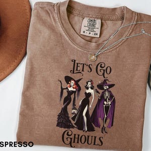 Puede incluir: Una camiseta marrón de manga corta con un estampado gráfico que muestra tres figuras con vestidos negros y un esqueleto con capa morada. El texto dice LET'S GO GHOULS. La camiseta tiene una etiqueta Comfort Colors y un collar de cadena plateada.