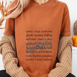 Puede incluir: Una camiseta de manga corta color óxido con una lista de palabras impresas en negro que incluyen spooky, creepy, mysterious, ghostly, haunted, chilling, enchanted, ghouls, candles, spiders, thrilling, skulls, supernatural, scares, bats, boo, graves, jackolanterns, witch, frightful, pumpkins, mystical, wicked, costumes y la palabra HALLOWEEN en una fuente estilizada.