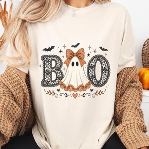 Puede incluir: Una camiseta color crema con un estampado gráfico de la palabra BOO. Las letras B y O están rellenas con un patrón de encaje negro. Entre ellas hay un fantasma blanco con un lazo naranja de lunares y una falda de encaje naranja a juego.