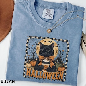 Puede incluir: Una camiseta de color azul vaquero con un gráfico de un gato negro con gafas de sol y pajarita, sosteniendo una taza con motivos de calabaza. El diseño incluye una luna llena, árboles desnudos, calabazas y la palabra HALLOWEEN en naranja.
