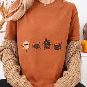 Puede incluir: Una camiseta de manga corta color óxido con un estampado gráfico de cuatro personajes de dibujos animados: una momia, una araña, una casa encantada y un caldero. Los personajes están dispuestos en una línea horizontal en el centro.