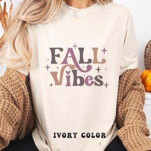 Puede incluir: Una camiseta de color marfil con el texto FALL VIBES en una fuente estilizada multicolor. El diseño incluye pequeños detalles de estrellas en tonos marrón, malva y beige. Las palabras IVORY COLOR están impresas en negro en la parte inferior.