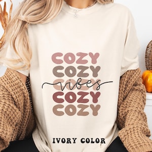 Puede incluir: Una camiseta de manga corta de color marfil con el texto "COZY" repetido cuatro veces en letras apiladas, redondeadas y de tonos apagados, con la palabra "vibes" escrita en cursiva negra en el centro. El texto "IVORY COLOR" aparece debajo.