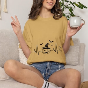 Puede incluir: Una camiseta de color amarillo mostaza con un estampado gráfico negro que muestra una línea de latido, una calabaza con sombrero de bruja, pequeños murciélagos volando y un símbolo de corazón. Tiene mangas cortas y cuello redondo.