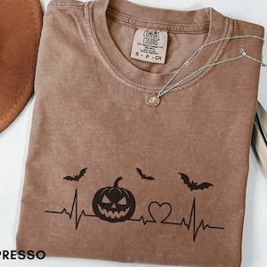 May include: An espresso-coloured t-shirt featuring a black graphic of a heartbeat line, a jack-o'-lantern, a heart, and two flying bats. The shirt has a crew neckline and a Comfort Colors brand label inside the collar. A silver necklace rests on top.