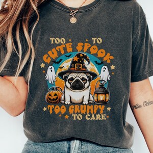 Puede incluir: Una camiseta gris carbón con un gráfico de un pug con sombrero de bruja, flanqueado por dos fantasmas, una calabaza y un farol. El texto "TOO CUTE TO SPOOK" y "TOO GRUMPY TO CARE" aparece en letras naranjas sobre un fondo de luna llena.