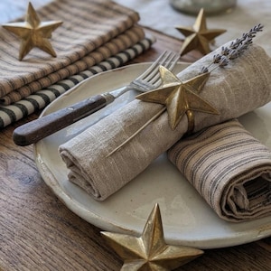 Peut inclure: Mise en place rustique avec une assiette blanche, des serviettes en lin roulées et une fourchette avec un manche en bois. Des ronds de serviette en forme d'étoile dorée et des étoiles décoratives ajoutent une touche d'élégance. Les serviettes ont un motif à rayures.