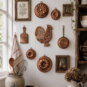 May include: A collection of copper kitchenware including various fluted moulds, a rooster-shaped wall hanging, a small skillet, and a wooden cutting board displayed on a white panelled wall alongside framed botanical and still life art prints.