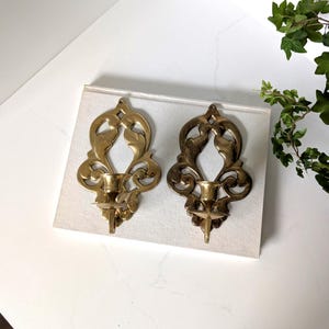 May include: A pair of matching gold-toned metal wall sconces featuring an openwork scroll design with a central candle cup. Each sconce has a symmetrical, elongated shape with a small hole at the top for mounting, showcasing a polished finish.