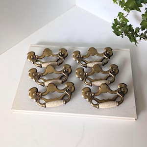 Peut inclure: Un ensemble de six poignées de tiroir en laiton dotées d'une structure métallique incurvée et ornée, avec un insert central en céramique blanc cassé moucheté. Chaque poignée présente un design symétrique aux extrémités arrondies et dorées.