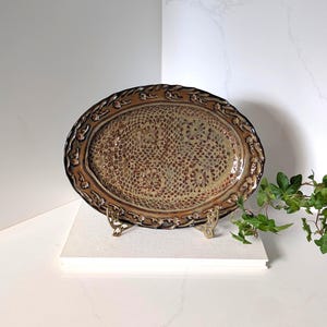 May include: An oval ceramic platter featuring a brown and tan mottled glaze with a textured, lace-like pattern across the centre. The rim is decorated with a raised, repeating floral motif in dark brown, supported by a gold-toned metal stand.