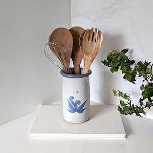 May include: A cylindrical ceramic utensil holder with a speckled off-white finish, a blue rim, and a blue floral design. The holder contains a metal wire whisk and three wooden kitchen utensils, including a solid spoon and a slotted salad fork.