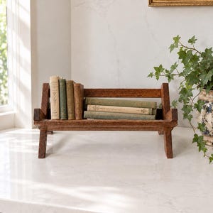 Peut inclure: Un petit porte-livres rectangulaire en bois avec une finition brun foncé et quatre pieds. Le support contient quatre livres anciens disposés verticalement sur la gauche et trois livres anciens empilés horizontalement sur la droite, aux tons vert et beige.