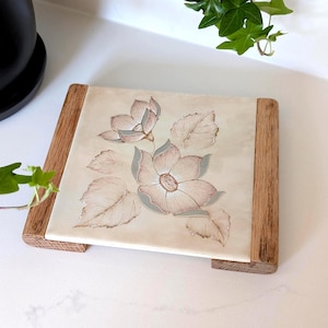 Peut inclure: Un dessous de plat rectangulaire composé d'un carreau en céramique crème orné d'une illustration florale délicate dans des tons rose poudré et vert sauge. Le carreau est encadré sur deux côtés par des lattes en bois clair au grain visible.
