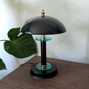 May include: A table lamp featuring a glossy black dome-shaped shade with a metallic finial at the top. The central column is black with two translucent teal glass circular accents, supported by a round black base and a black power cord.