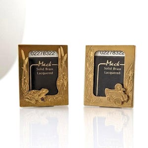 May include: Two rectangular solid brass picture frames with a lacquered finish. The left frame features a frog and cattails, while the right frame features a duck and floral motifs. Both frames contain a black insert with the text Mack Solid Brass Lacquered and the number 02278322.