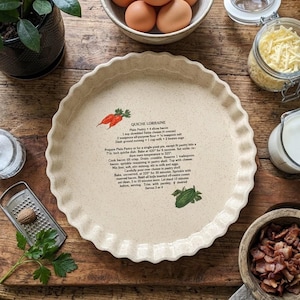 May include: A round, cream-coloured ceramic quiche dish with a fluted edge. The centre features a printed Quiche Lorraine recipe with instructions and ingredient lists, accompanied by small illustrations of carrots and a green vegetable.