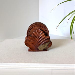 May include: A dark brown wooden coaster holder shaped like a large leaf, containing a set of circular wooden coasters. A small, metallic, gold-toned plaque shaped like the island of Cyprus is attached to the front of the leaf-shaped holder.