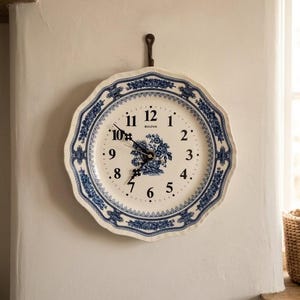 May include: A round wall clock shaped like a scalloped plate with a cream-coloured face and a blue floral border. The clock features black Arabic numerals, black hands, and the word Bulova printed in black text above a central blue floral motif.