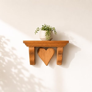 May include: A medium-toned wooden wall shelf featuring a carved heart shape centred between two decorative corbel supports. A small, light-coloured ceramic pot containing a trailing green plant sits on the flat top surface of the shelf.